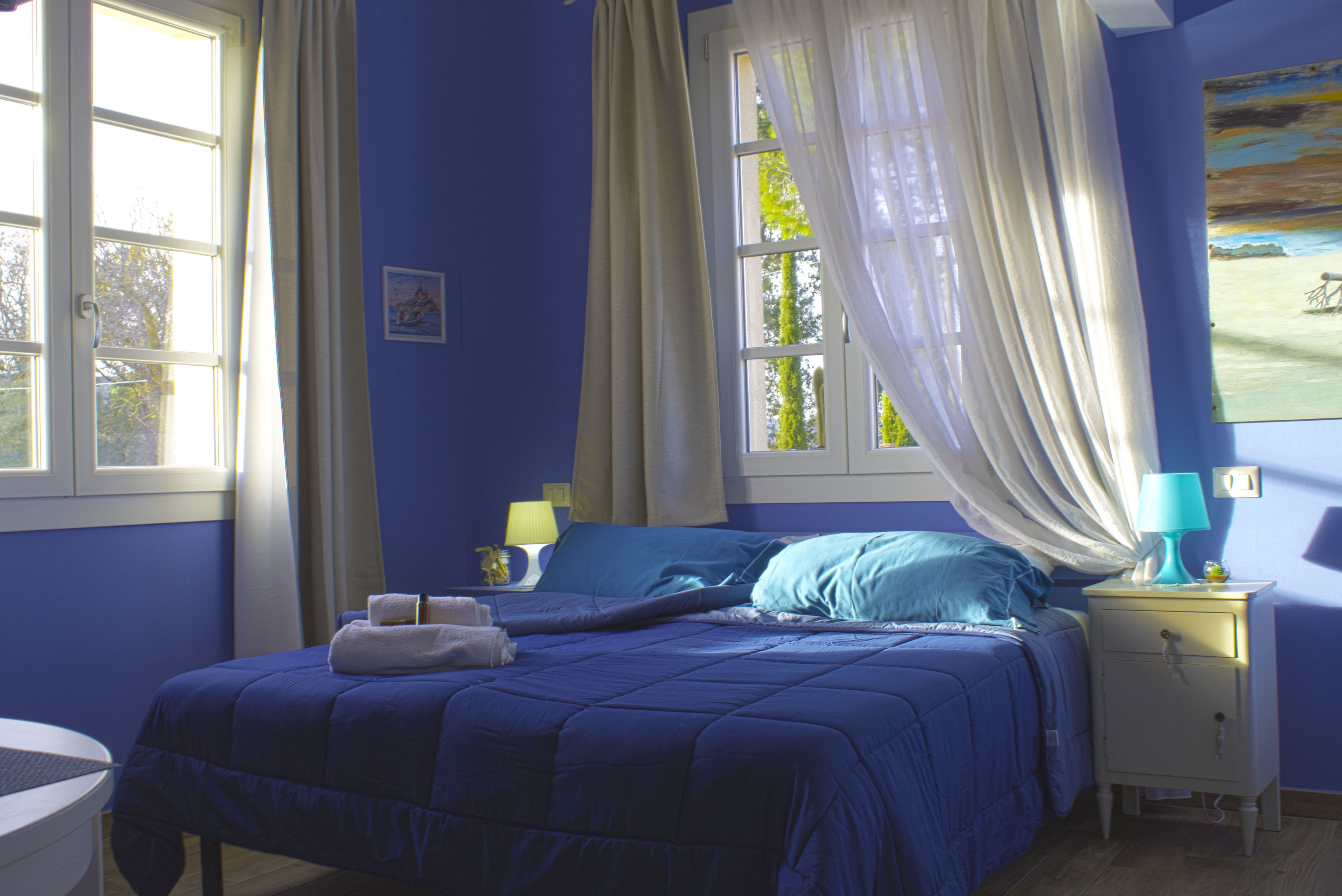 Una camera da letto con un letto, un comodino, una finestra con tende e un quadro appeso al muro.