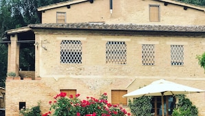 A building with a stone facade, multiple windows with metal grilles, and a large potted plant with red flowers in the foreground. - Small Apartment With Independent Access  In Beautiful Tuscan Farmhouse (Castelnuovo Berardenga)