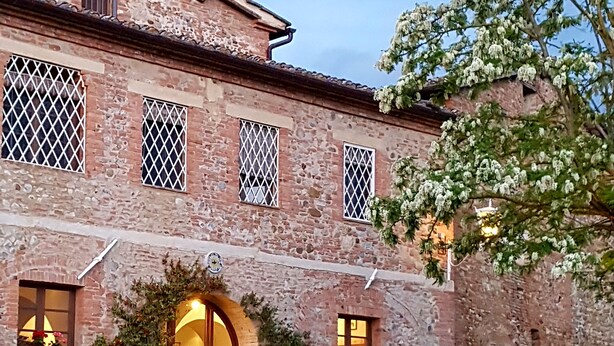 A rustic stone building with a central arched entrance, multiple windows with metal grilles, and outdoor seating area with tables and chairs. - Small Apartment With Independent Access  In Beautiful Tuscan Farmhouse (Castelnuovo Berardenga)