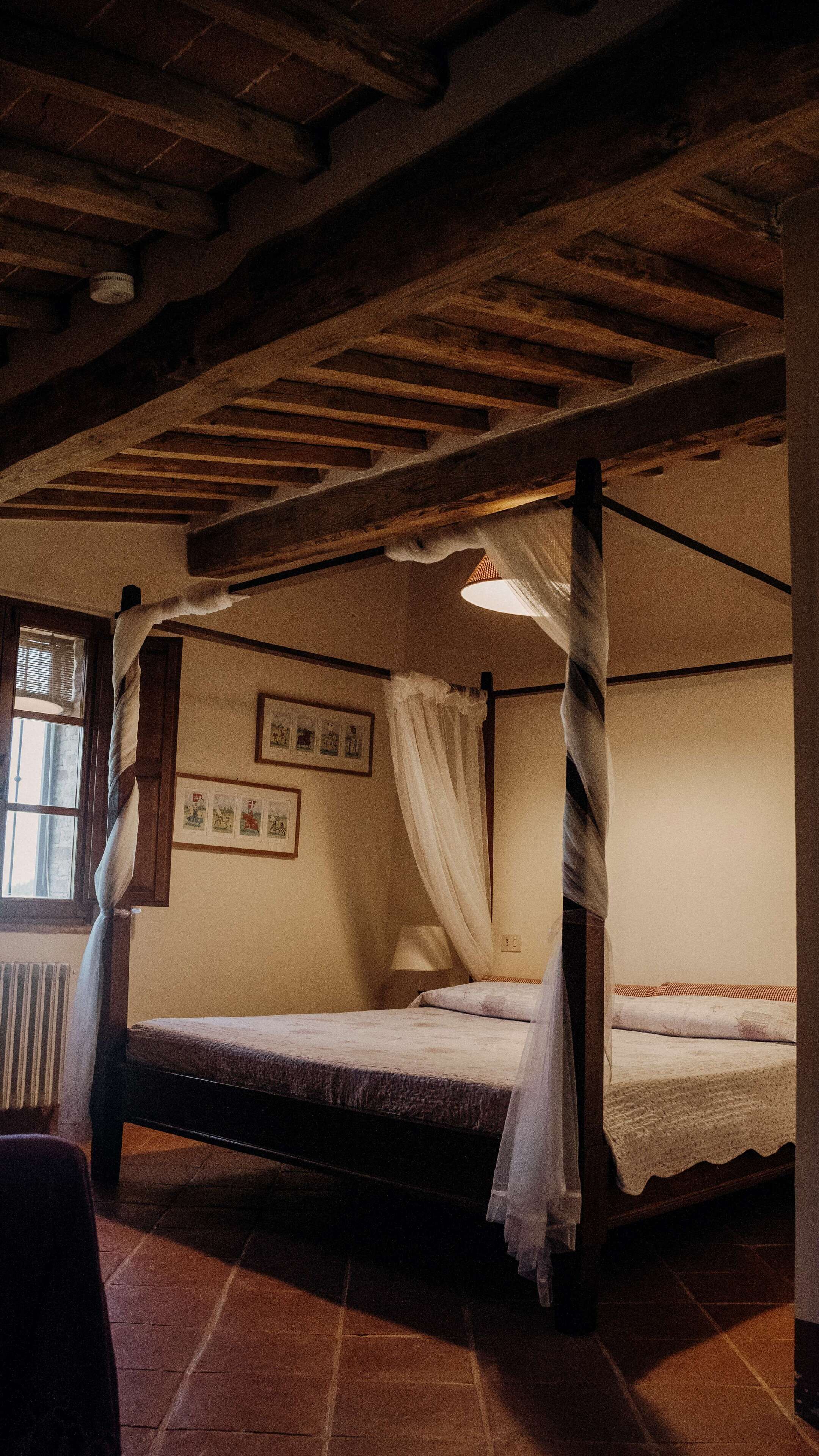 A rustic room with a four-poster bed, wooden beams on the ceiling, and a window with curtains.