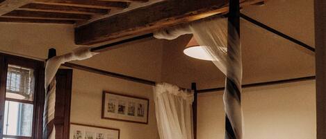 A rustic room with a four-poster bed, wooden beams on the ceiling, and a window with curtains.