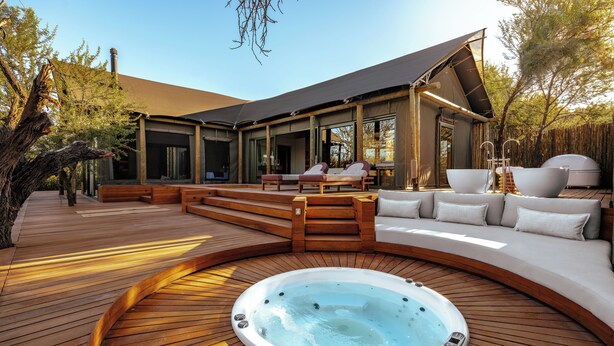 Un jacuzzi rodeado de una terraza de madera, un área de estar cubierta y un edificio moderno con grandes ventanales.