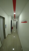 Un pasillo largo y estrecho con paredes blancas y techo rojo. Se observa un extintor de incendios en la pared, además de un cartel con números de habitación.