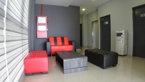 Un moderno vestíbulo de hotel con un sofá, reposapiés y una pequeña mesa. Se observa una unidad de aire acondicionado instalada en la pared y una puerta con la etiqueta 'SURAU'.