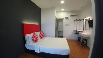 Habitación de hotel con cama, cabecera roja, colchón blanco, piso de madera, baño con lavamanos y espejo, y unidad de aire acondicionado.