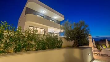 A modern two-story house with a balcony, outdoor seating area, and a well-lit garden.