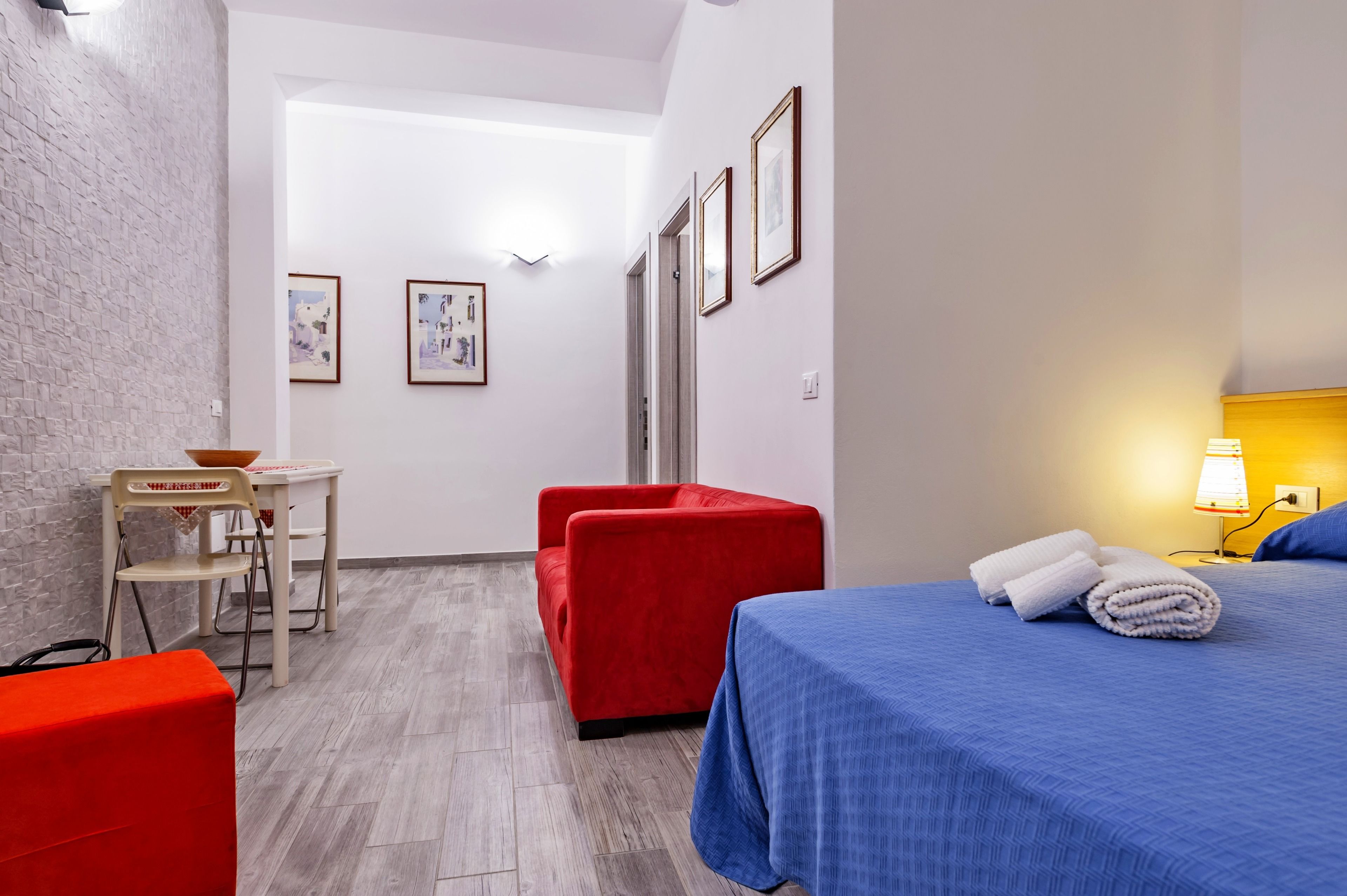 Une chambre d’hôtel avec un lit, une chaise rouge, une petite table avec un bol, et des tableaux encadrés accrochés au mur.