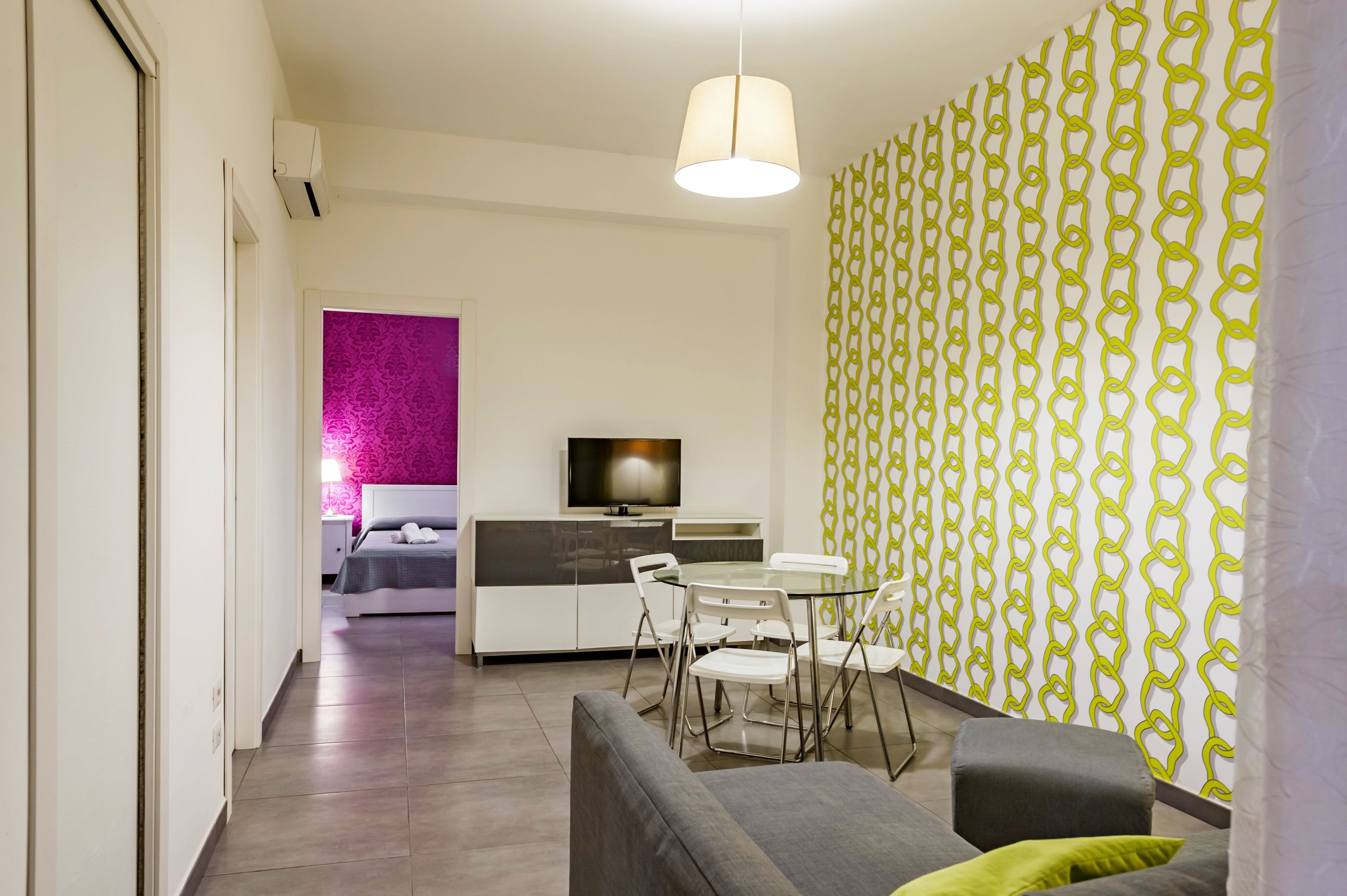 Un soggiorno moderno con un divano grigio, un tavolo da pranzo in vetro con sedie bianche, un televisore a schermo piatto su un mobile bianco e una parete decorata con un motivo.