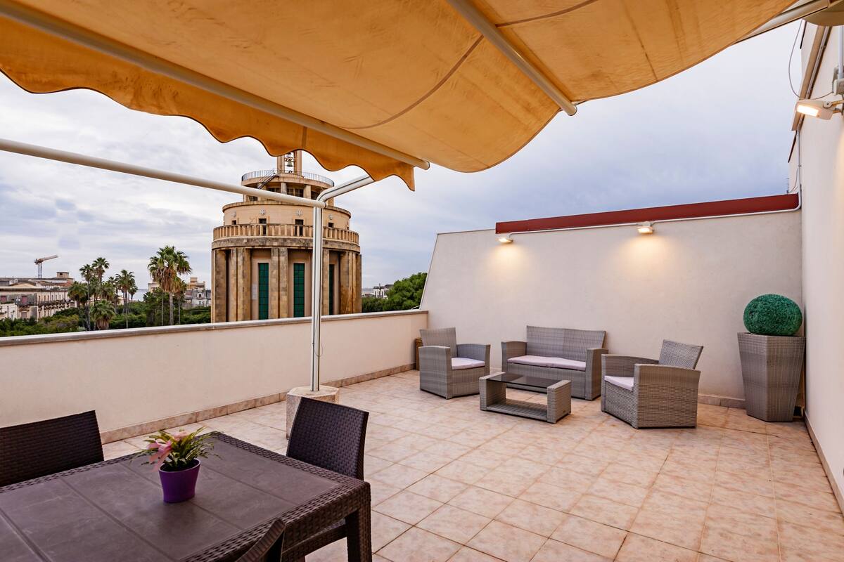 A rooftop terrace with wicker furniture, a table, and a potted plant.