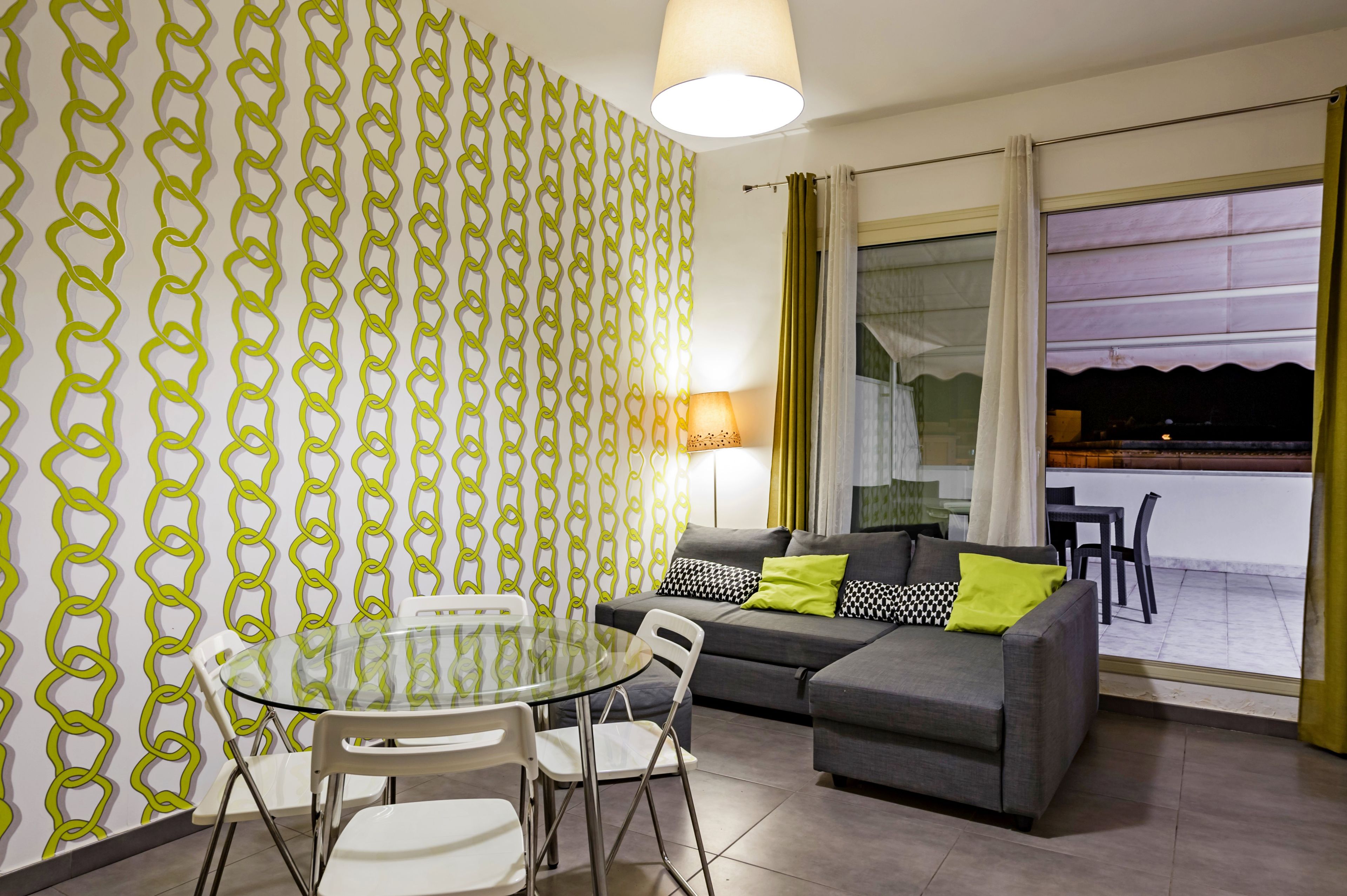 A modern living room with a grey sofa, a glass table, and white chairs. A patterned wallpaper and a large window with curtains are visible.