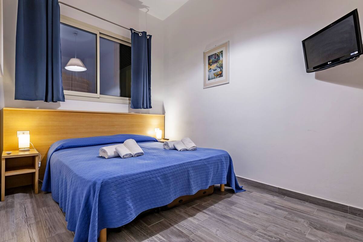 A neatly made bed with blue bedding, a bedside lamp, and a window with curtains.