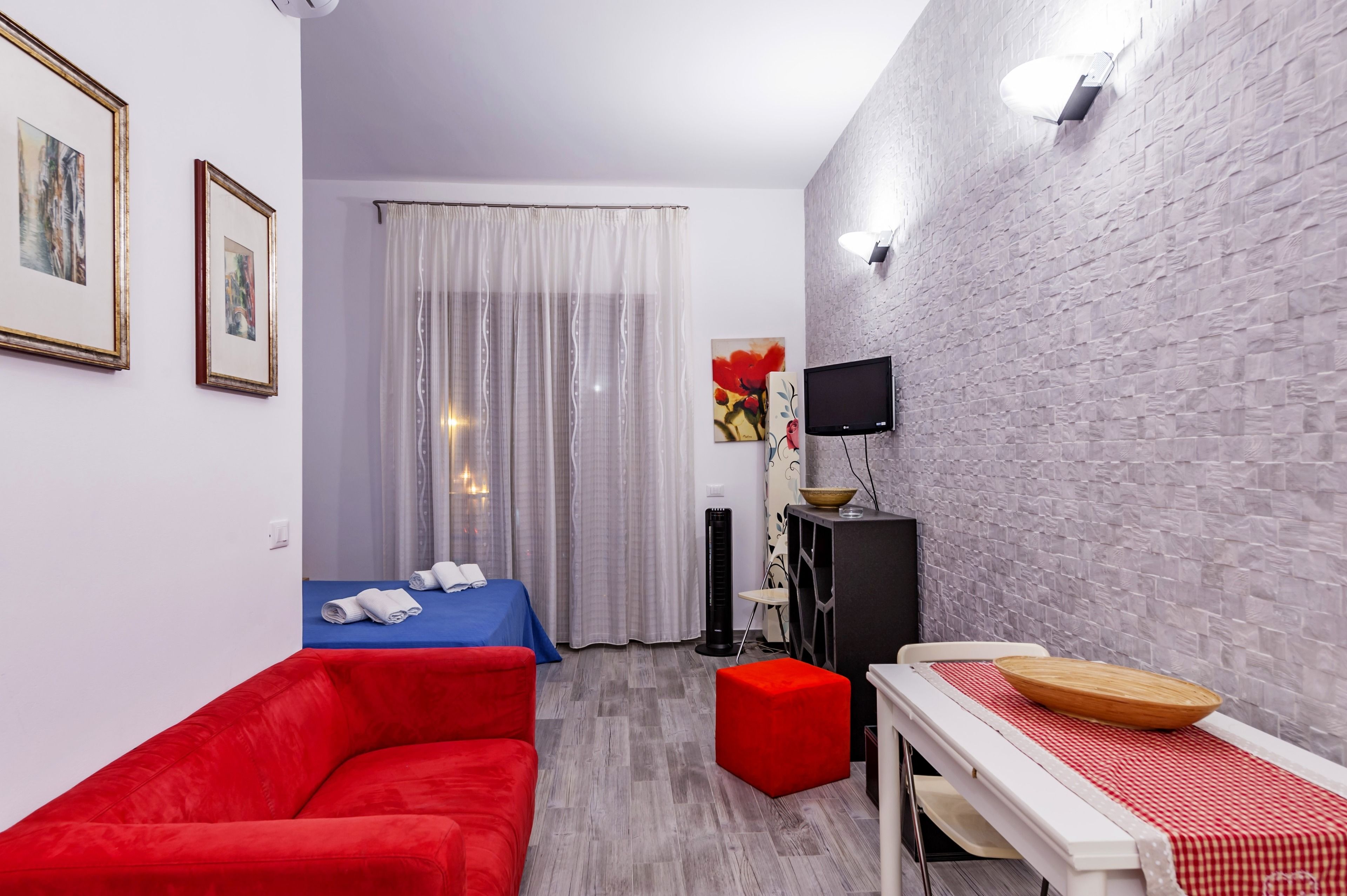 Habitación de hotel compacta con sofá rojo, cama azul, pequeña mesa de comedor con mantel a cuadros rojo y blanco, televisor de pantalla plana y pared decorativa con textura.