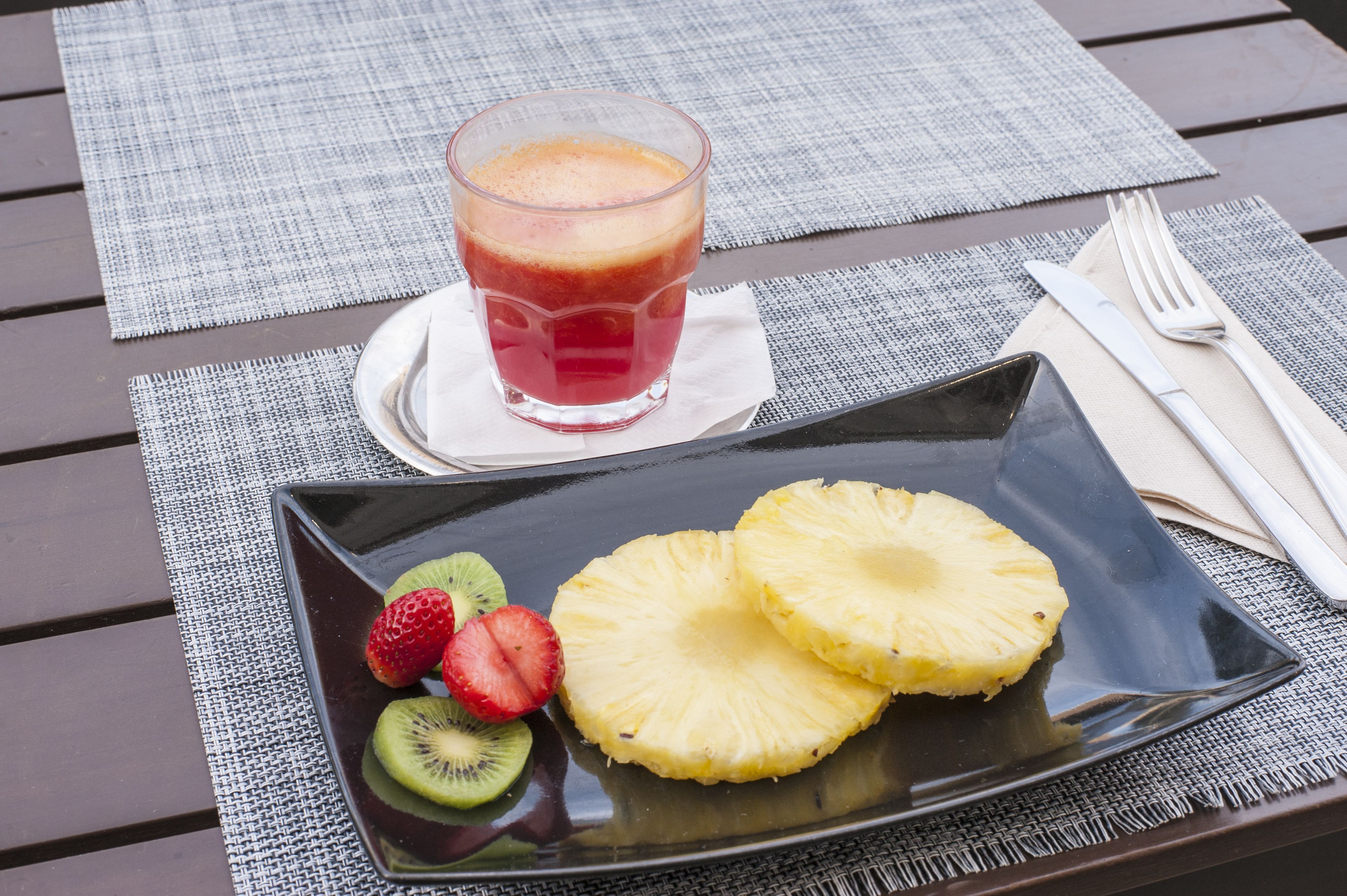 Comida con un vaso de jugo, rodajas de piña y kiwi en un plato, sobre una mesa con posavasos.