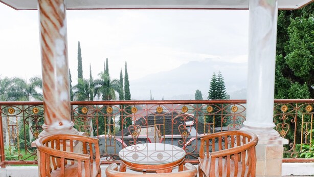 Un balcón con mesa y sillas de madera, barandilla metálica decorativa y vista a árboles y montañas al fondo.