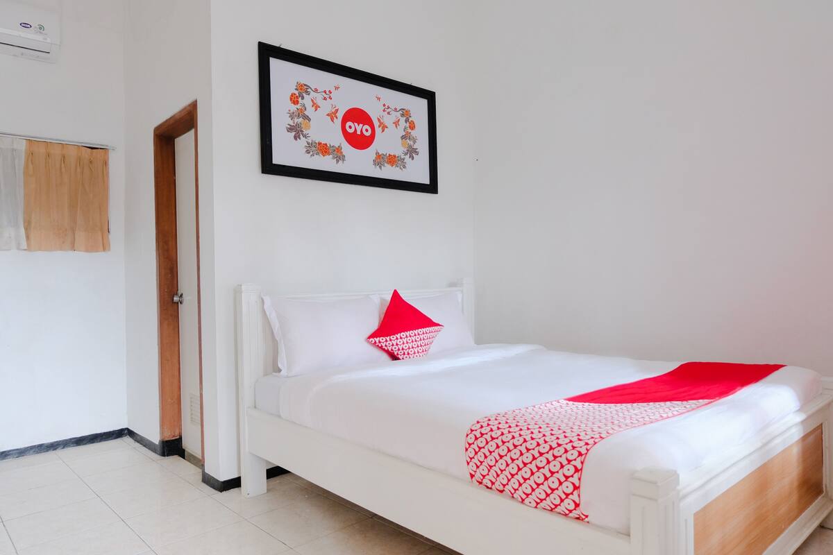A neatly made bed with white bedding and a red pillow, a framed picture on the wall, and a wooden door.