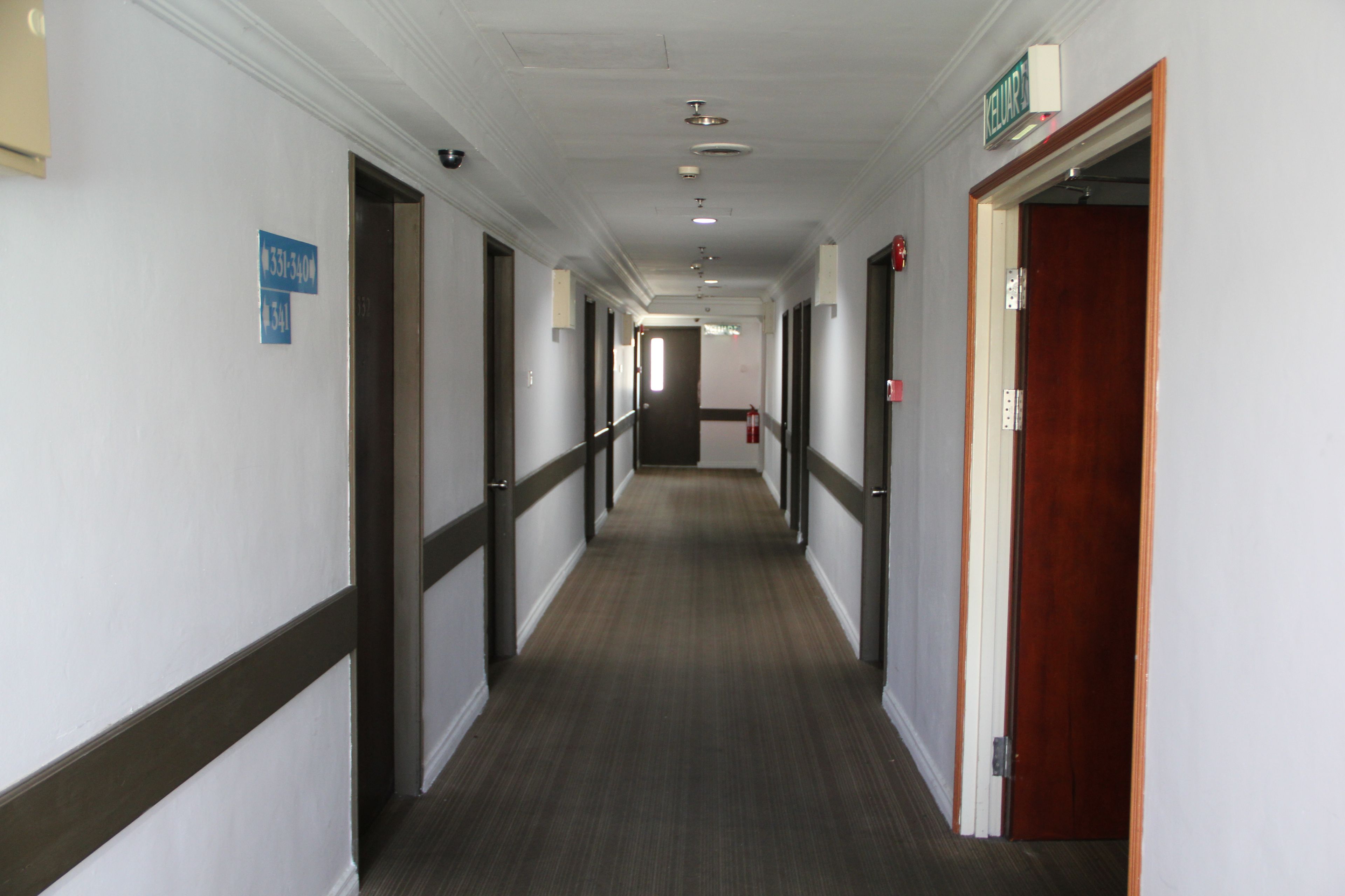 A long, narrow hallway with doors on both sides, a fire extinguisher on the right wall, and a sign indicating the direction to the elevator.