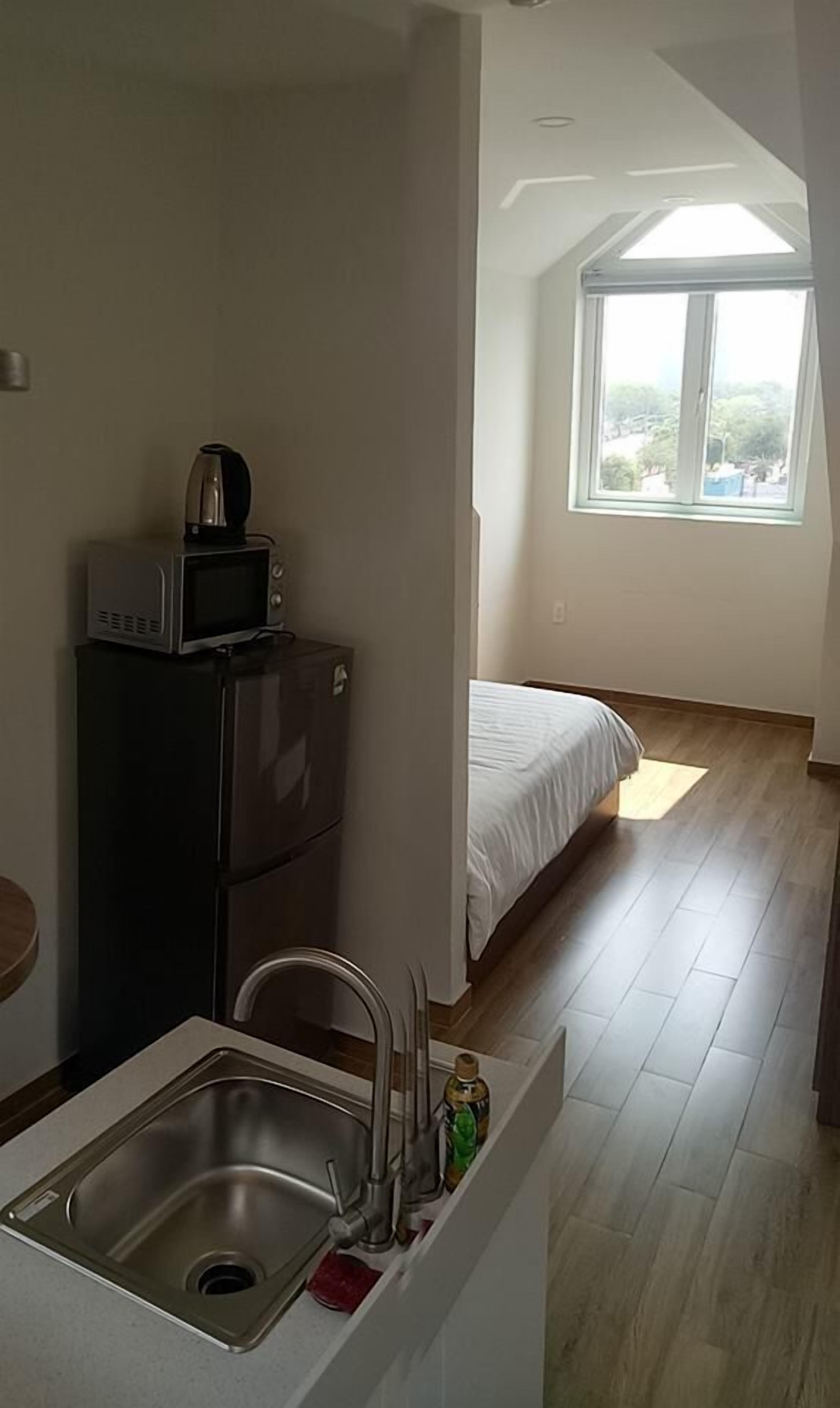 A compact kitchen with a microwave, sink, and a small refrigerator. A bed is visible in the adjacent room with a large window providing natural light.