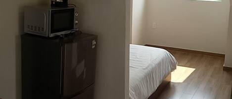 A compact kitchen with a microwave, sink, and a small refrigerator. A bed is visible in the adjacent room with a large window providing natural light.