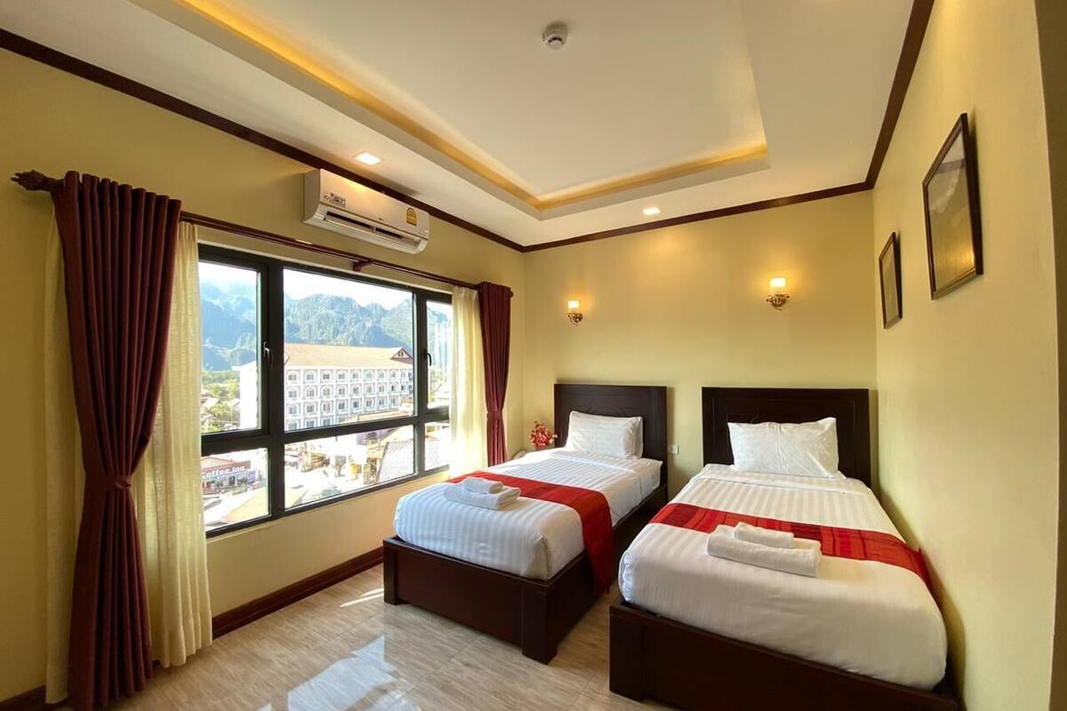 Habitación de hotel con dos camas, un ventanal con vistas a edificios, un equipo de aire acondicionado y una lámpara en el techo.