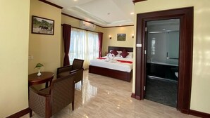 Une chambre d’hôtel avec un lit, un bureau avec une chaise, une petite table avec une plante, et une salle de bain visible à travers une porte ouverte.