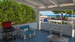 A covered outdoor patio with a barbecue grill, a blue cushioned seating area, and a view of the sea.