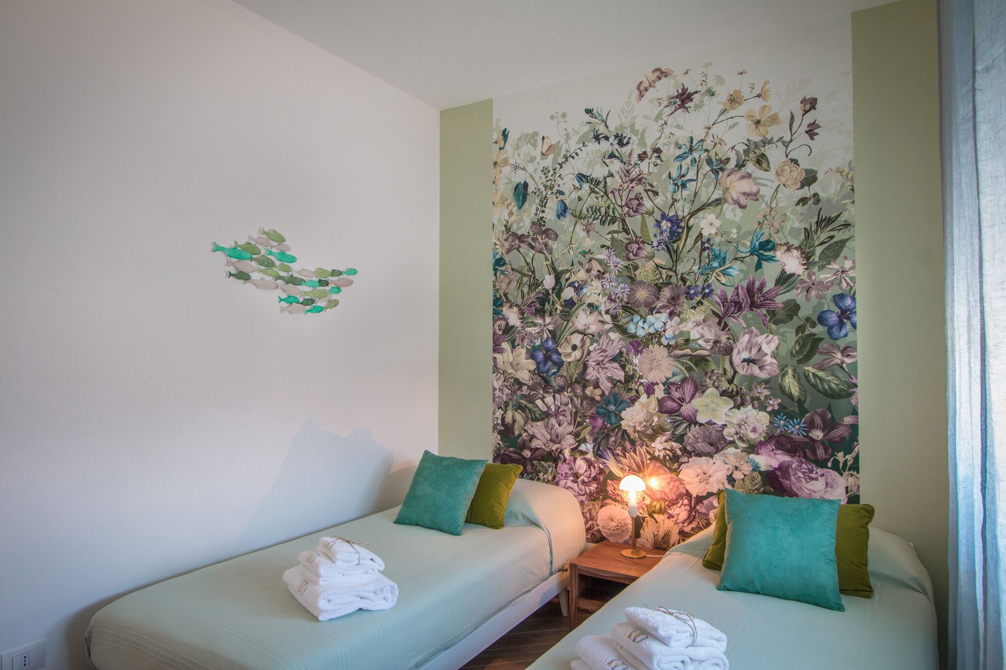 Une chambre d’hôtel avec deux lits, chacun agrémenté d’un oreiller vert, d’un papier peint à motifs floraux et d’une lampe de chevet.
