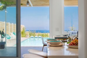 Comedor con vista a una piscina y al océano. Hay sillas blancas, una mesa con platos y vasos, y un bol de fruta.