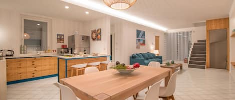 A modern kitchen with a wooden island, white chairs, and a dining table with a bowl of fruit.
