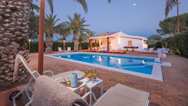 Piscina con sillas de descanso y una mesa, rodeada de palmeras y una casa.