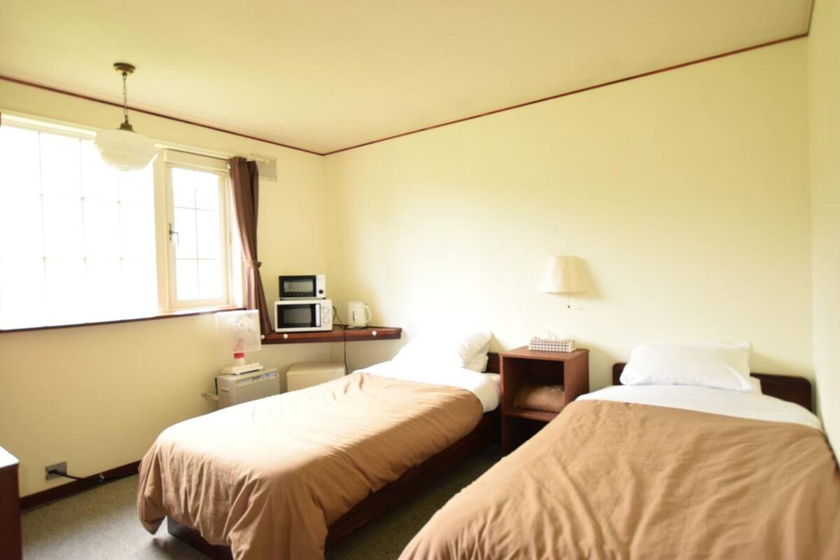 A small, simple room with two beds, a desk with a microwave, a lamp, and a window with curtains.