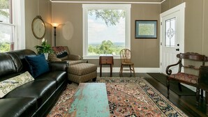 Un salon chaleureux avec un canapé en cuir noir, un pouf à motifs et une grande fenêtre donnant sur un paysage verdoyant.