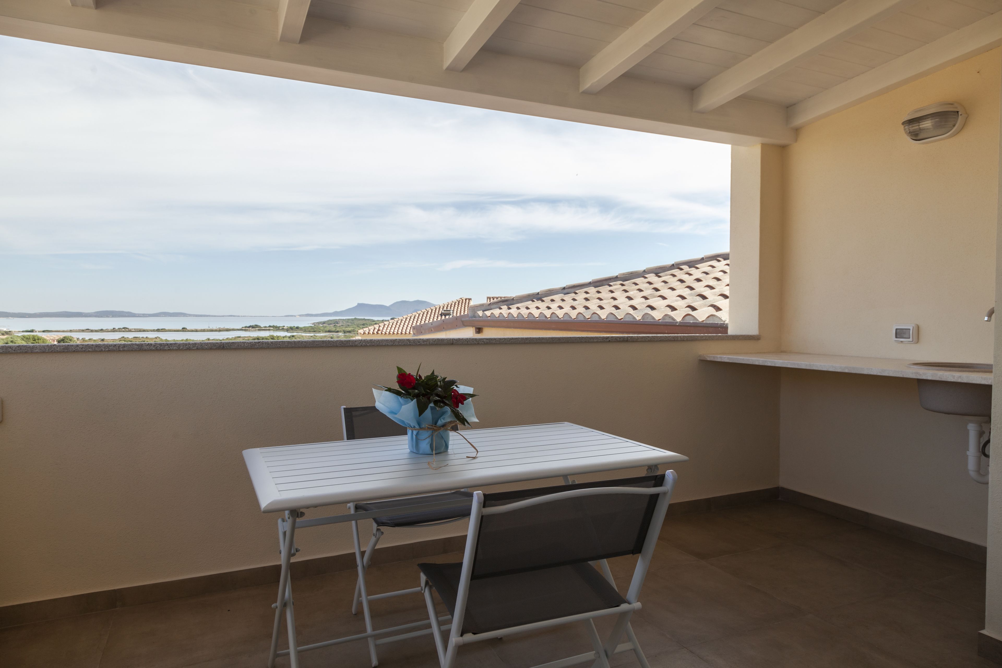 Un balcon avec une table et des chaises, un vase de fleurs et une vue sur la mer et les montagnes lointaines.