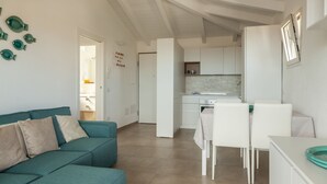 A modern living room with a teal sofa, a dining area with white chairs, and a kitchen with white cabinets. - Aria di Vacanza - Azalee (Olbia)