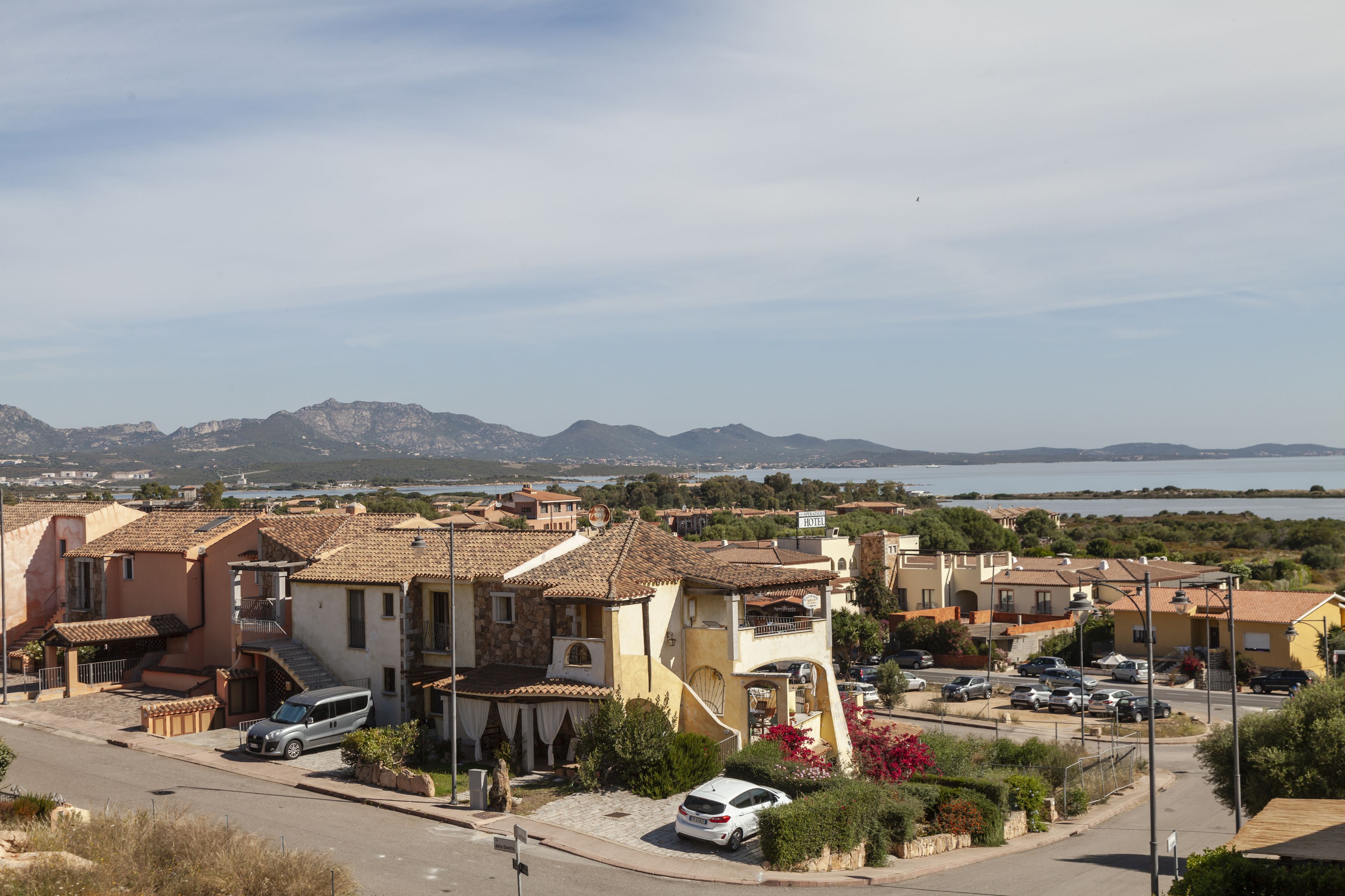 A coastal residential area with terracotta-roofed houses, a paved street, and a view of the sea and mountains in the distance.