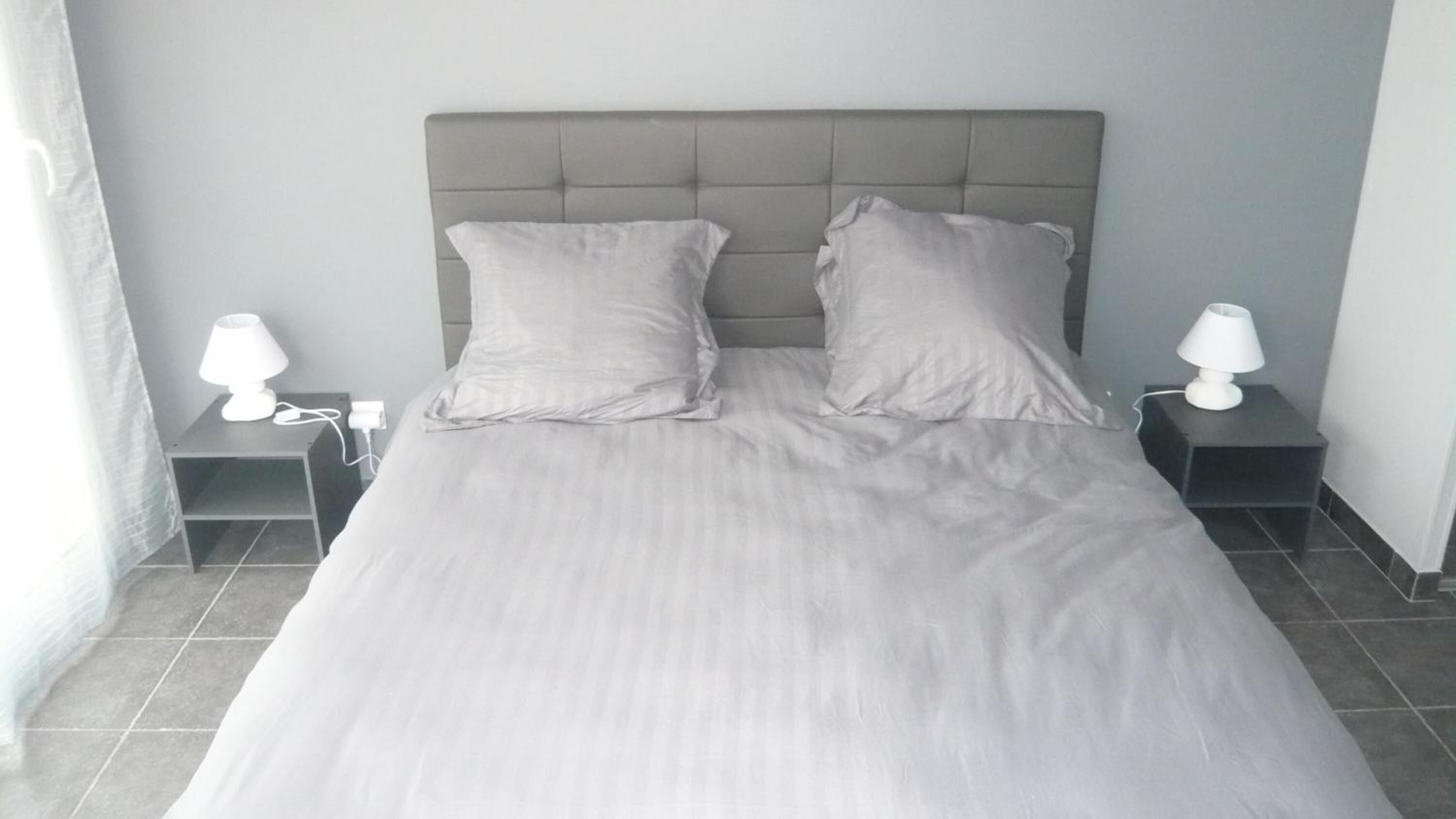 Een opgemaakt bed met twee kussens en een grijs hoofdeinde. Aan beide zijden van het bed staan nachtkastjes met lampen.