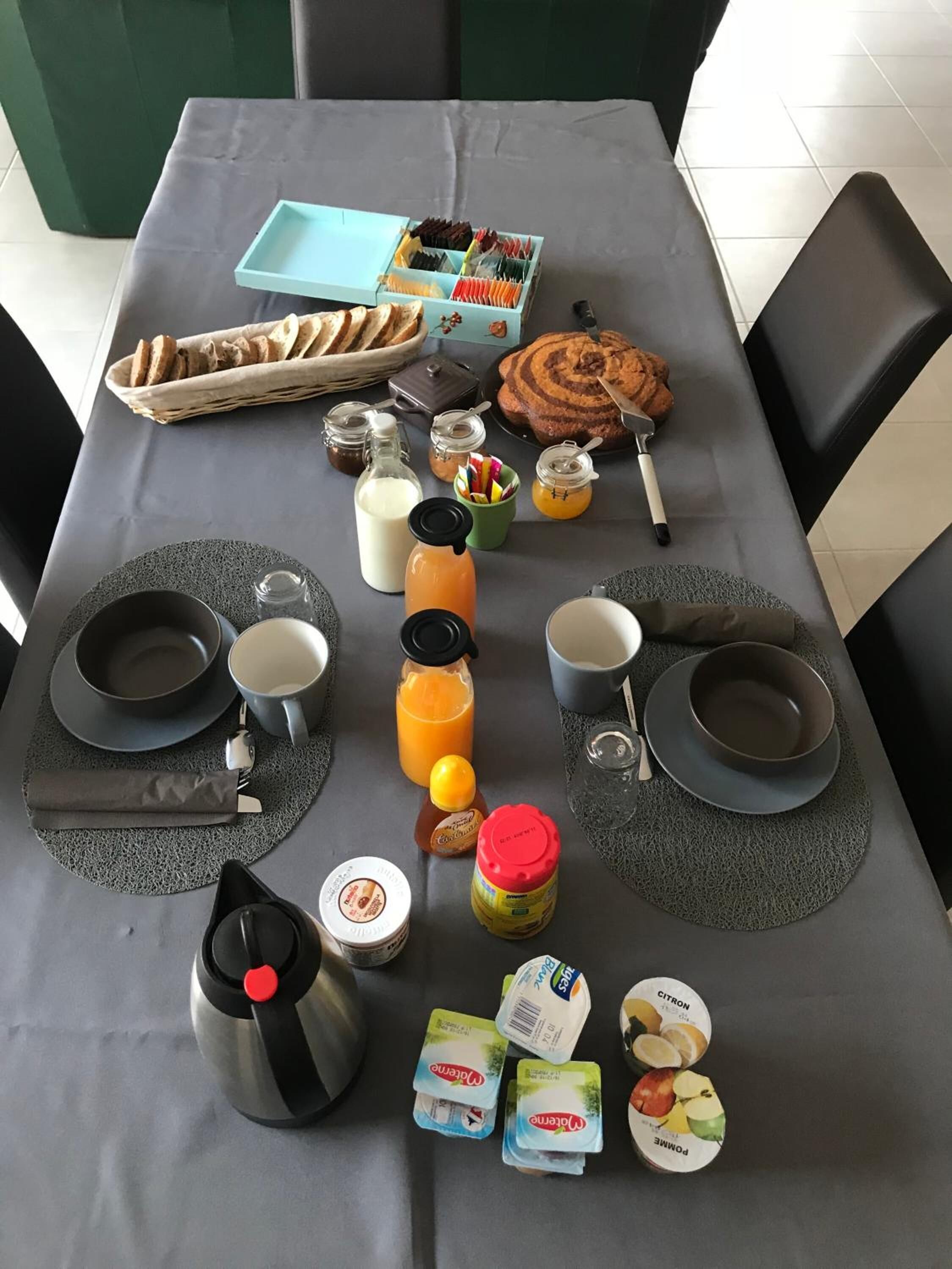 A breakfast table set with plates, bowls, cups, a coffee pot, and various food items including bread, pastries, and condiments.