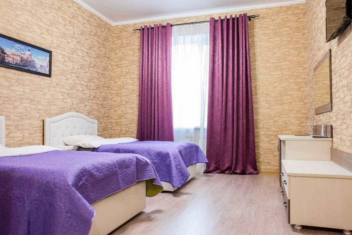 Habitación de hotel con dos camas, una pared de madera, un ventanal con cortinas moradas y un escritorio pequeño con espejo.