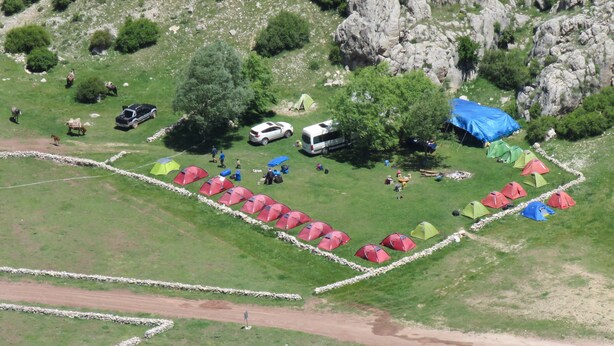 Um camping com barracas dispostas em fileiras, um caminho de terra e um morro rochoso ao fundo.