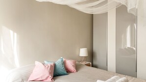 Cama bem arrumada com almofadas rosa e azul, luminária branca em uma mesa de cabeceira e um dossel branco sobre a cama.