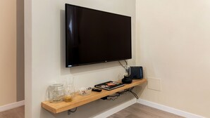 Un téléviseur fixé au mur, au-dessus d’une étagère en bois, avec une télécommande, un récipient en verre et un livre.