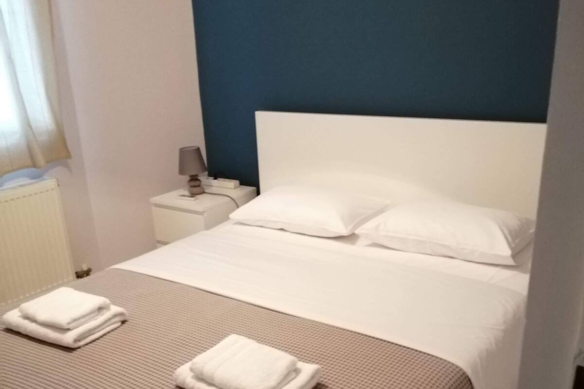 A neatly made bed with white linens and two folded towels, a bedside table with a lamp, and a wall with a small artwork.