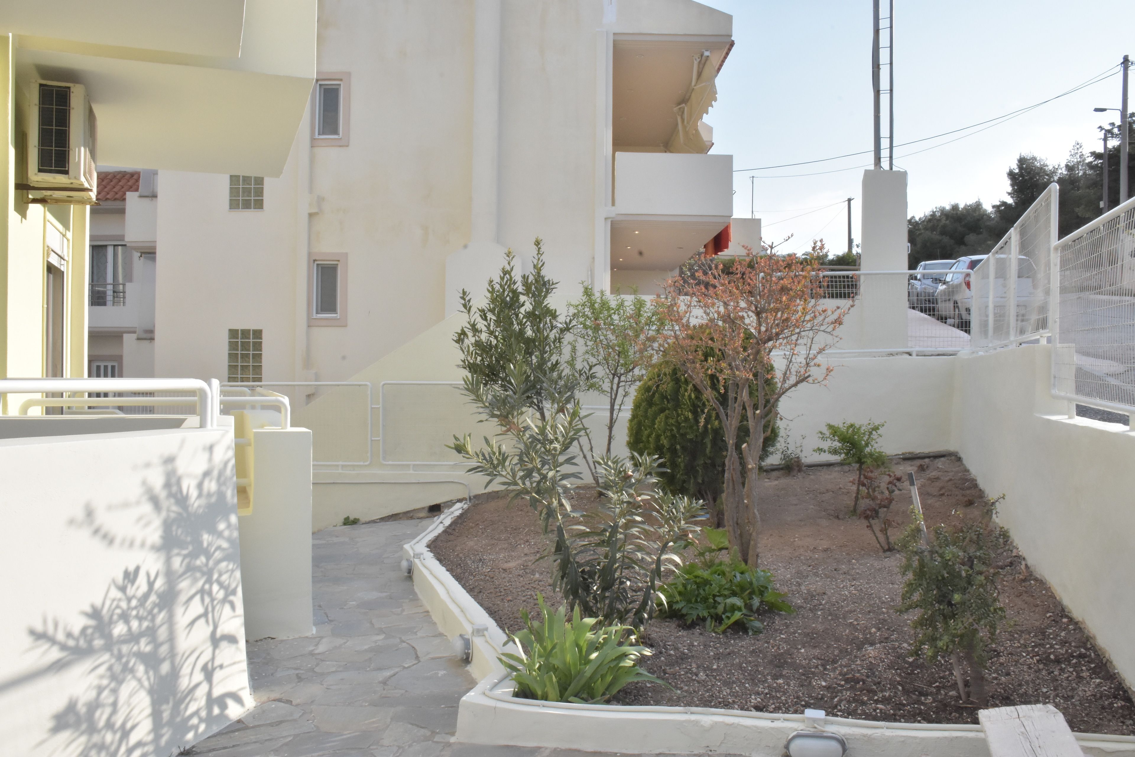 Un patio con un pequeño jardín, un edificio blanco con balcones y un automóvil estacionado al fondo.