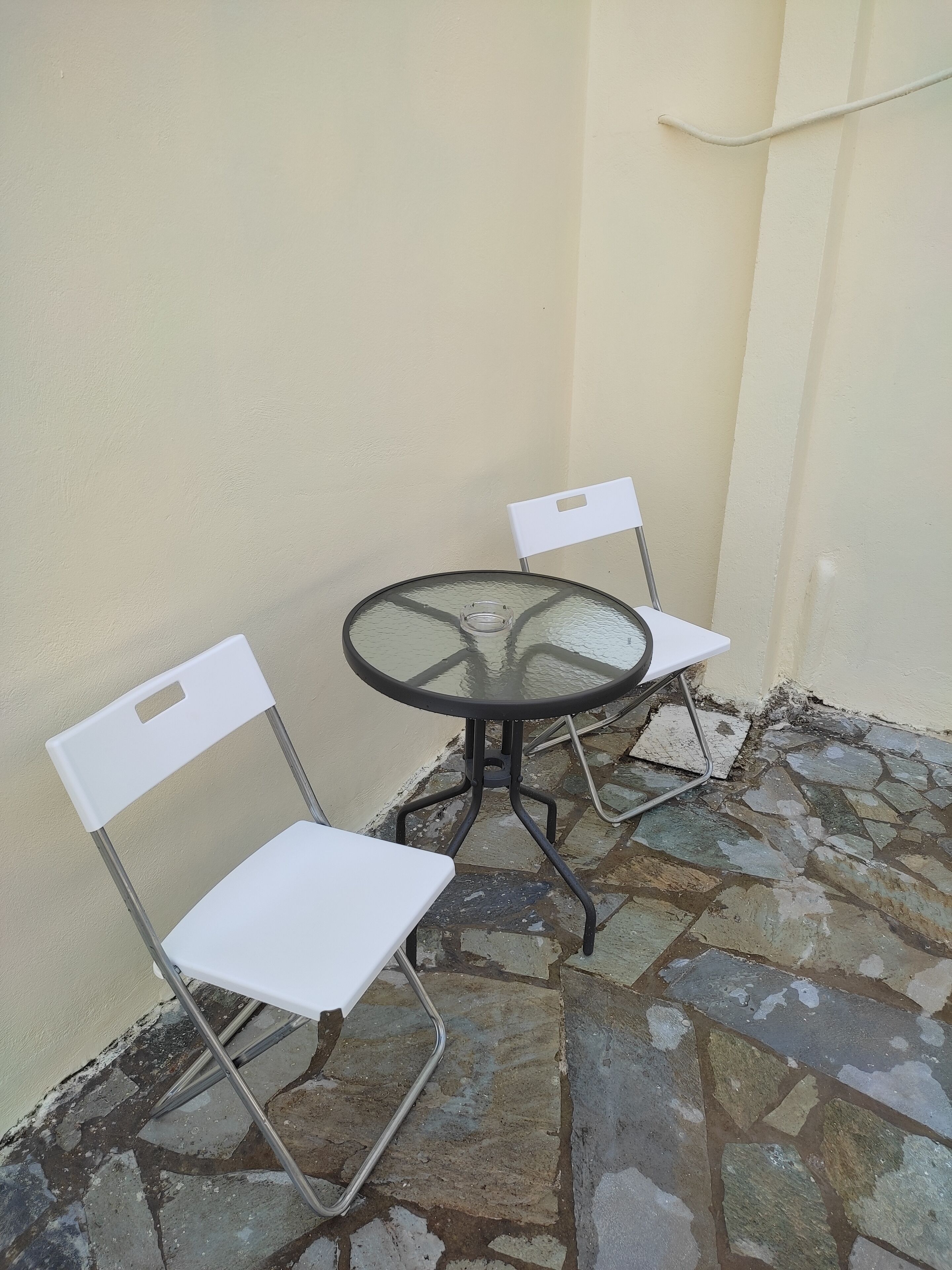 Une petite table d’extérieur avec un plateau en verre et un cadre en métal, accompagnée de deux chaises blanches aux pieds en métal, est placée sur une terrasse carrelée en pierre, adossée à un mur sobre.
