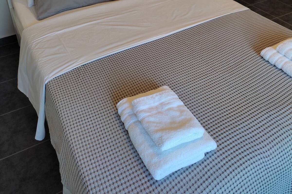 A neatly made bed with a grey patterned bedspread and two grey pillows, a white bedside table with a remote control, and a white towel folded on the bed.