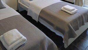 Ein Hotelzimmer mit zwei Betten, jeweils mit weißem Kopfteil, grau-weißer Bettwäsche und einer karierten Decke. An den Nachttischen stehen Lampen, Steckdosen und Fernbedienungen. Das Zimmer verfügt über ein Fenster mit beigen Vorhängen.