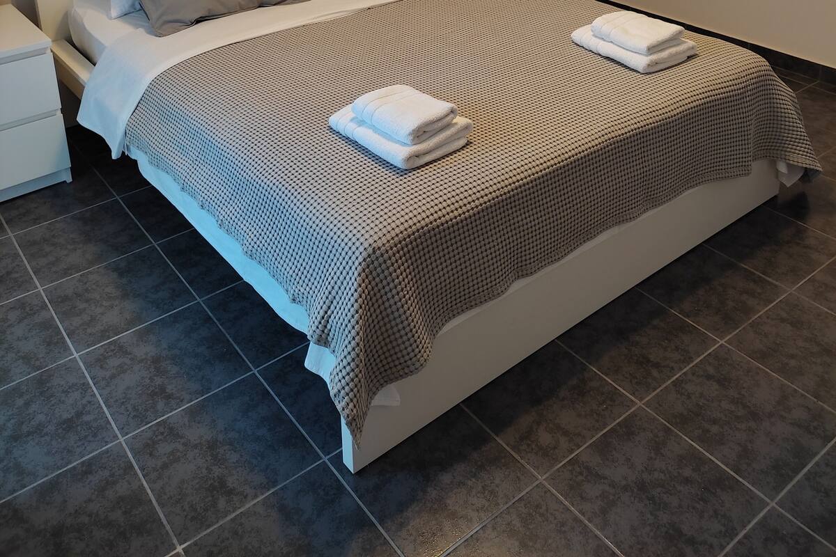 A neatly made bed with a checkered bedspread and two folded towels, a bedside lamp, and a nightstand with a remote control.