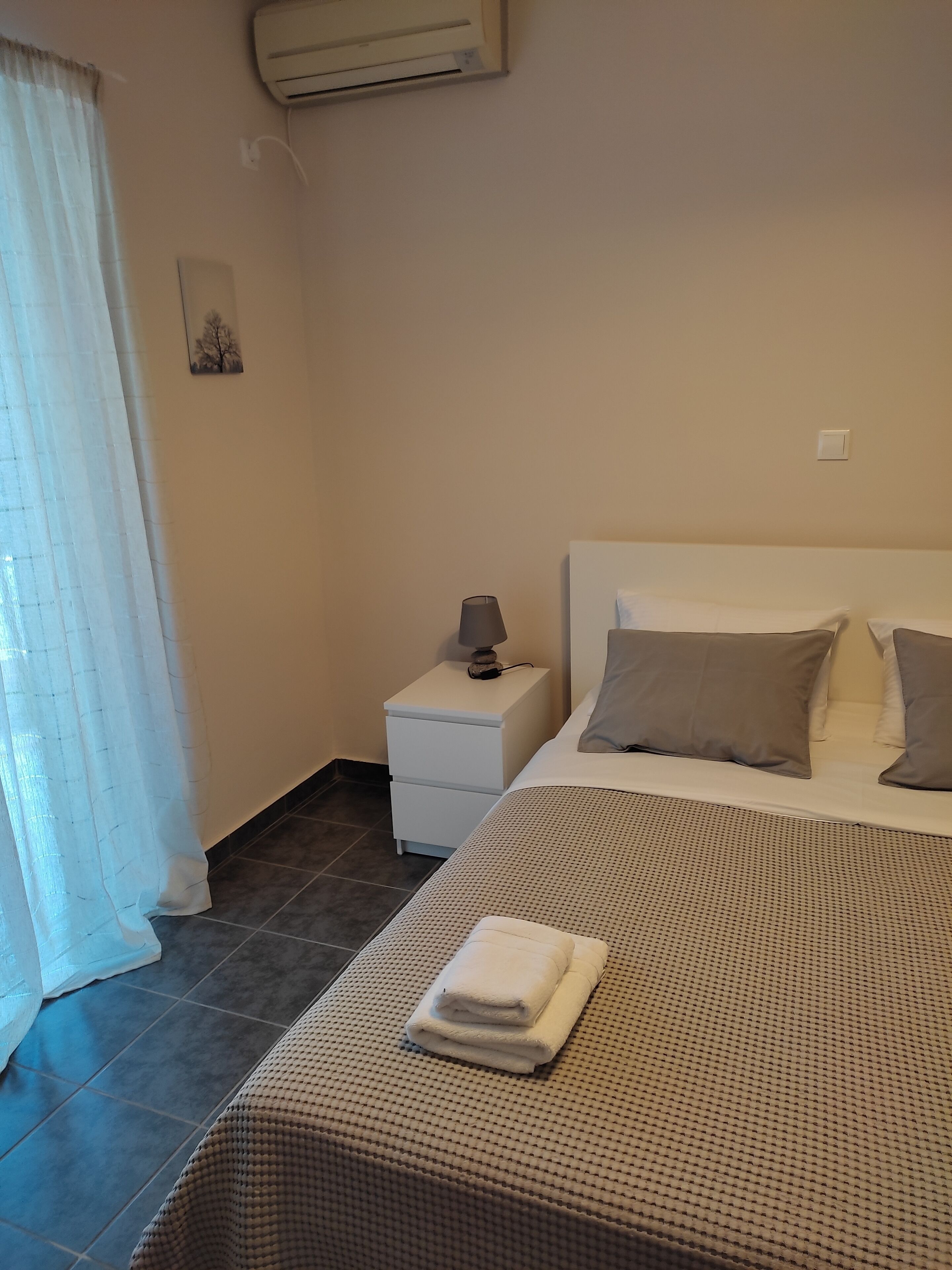 Quarto arrumado com cama, travesseiros, luminária de cabeceira, mesa de cabeceira e toalha dobrada sobre a cama.
