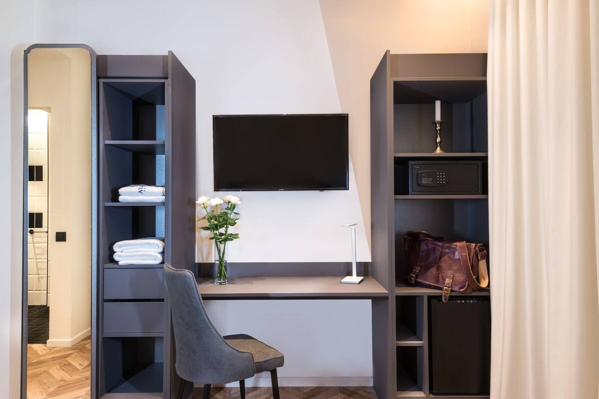 A modern hotel room with a desk, a flat-screen TV, a safe, and a vanity with a mirror.