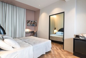 Quarto de hotel com cama, escrivaninha, cadeira, espelho e pequeno frigorífico.