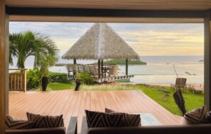 Eine Holzdielen-Terrasse mit einem strohgedeckten Gebäude mit Blick auf einen Strand.